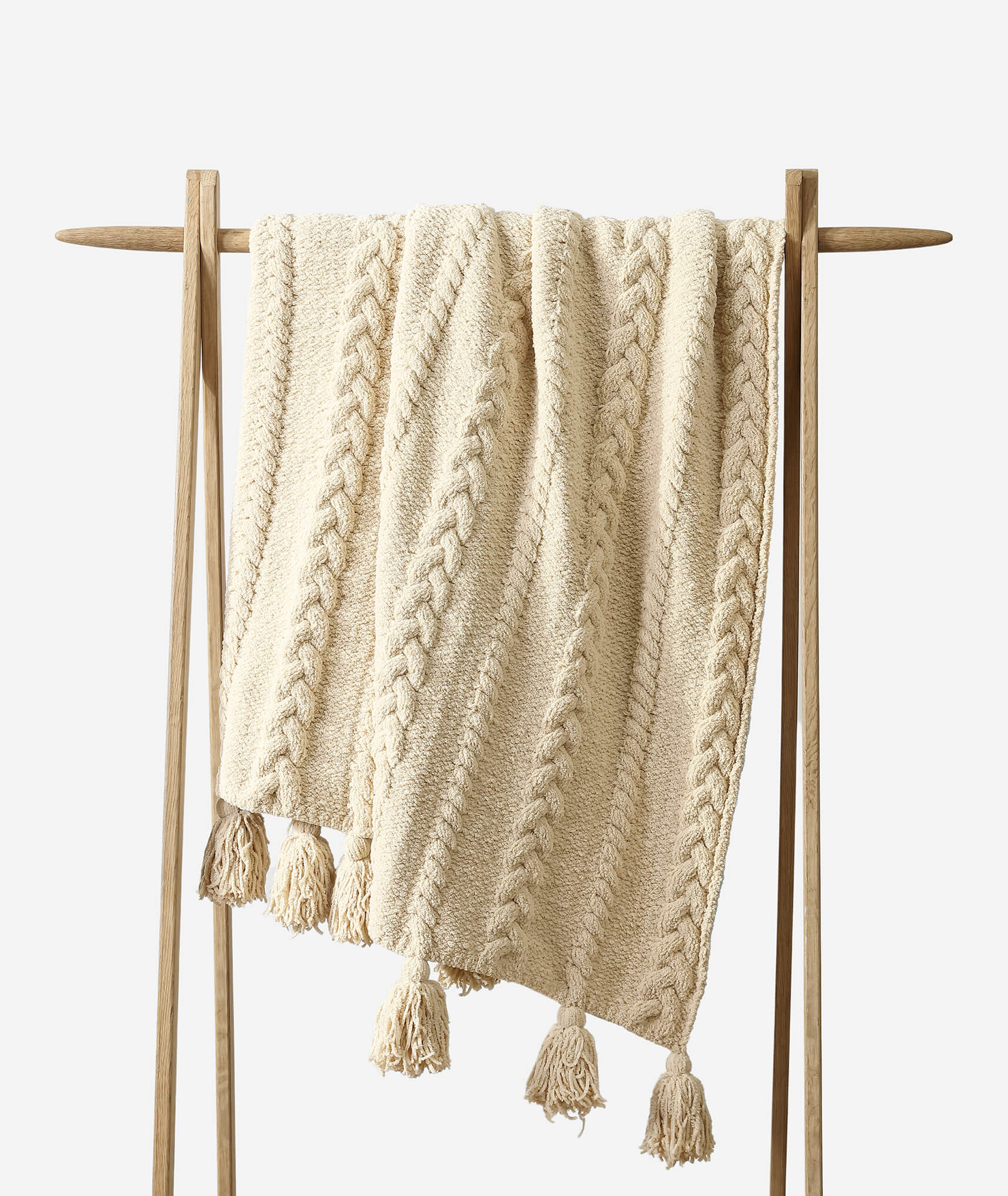 A cream-colored, cable-knit Braided Pom Pom Throw by Sunday Citizen with tasseled ends is draped over a minimalist wooden stand against a white background. Crafted from breathable, cozy fabric, the throw features a textured pattern with chunky braided designs and is OEKO-TEX® certified for added peace of mind.