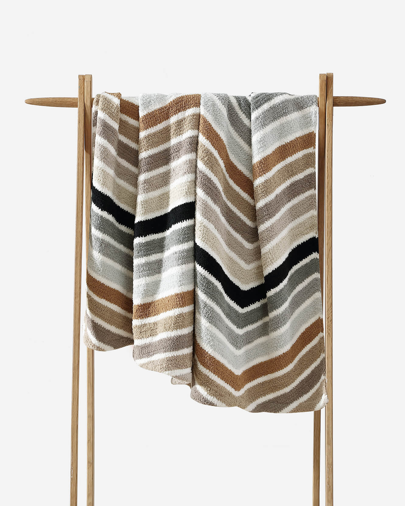 A cozy blanket draped over a wooden rack. This Cusco Lightweight Throw by Sunday Citizen features a striped zigzag pattern in shades of brown, beige, gray, and black, creating a warm and inviting look. The snug fabric is also machine washable for easy care.