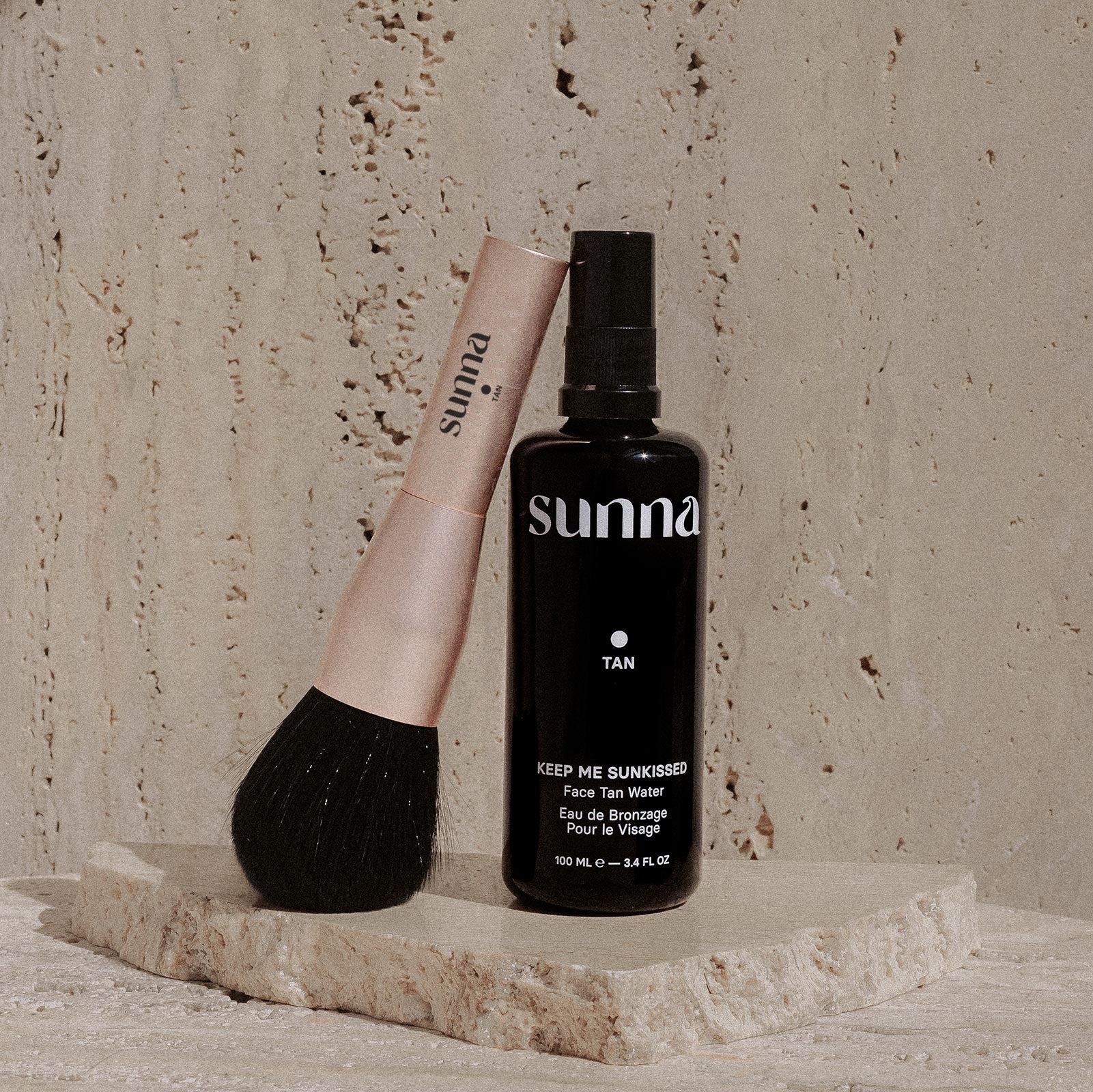 An image of Sunna tanning products. The set includes a black spray bottle labeled "Face Tan Water + Brush by Sunna" with anti-ageing ingredients and a makeup brush with a black bristle head and a gold handle. Both are placed on a beige stone slab against a matching textured background, promising glowing skin.