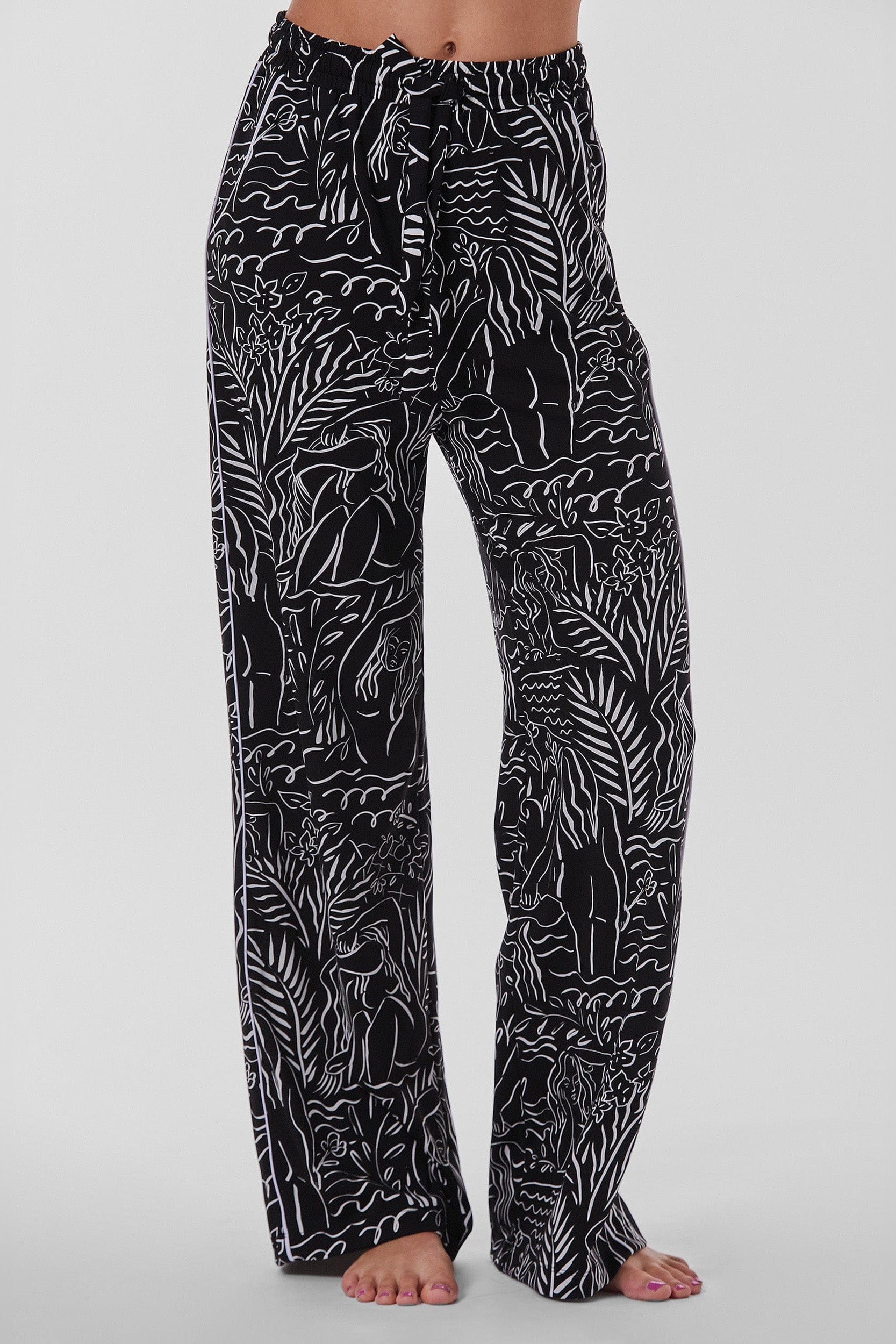 A person is shown from the waist down, barefoot on a plain white background, wearing the Venus Pajama Soft Long Pant. These high-waisted, flowy pants feature an abstract jungle-themed pattern with plants and animals in black and white. The soft material and tied waistband embody a distinct Venus Print allure.