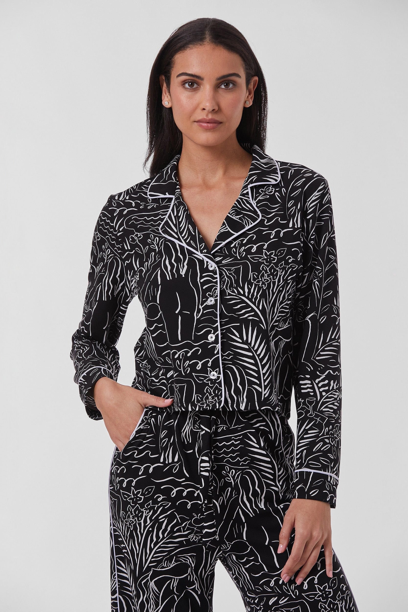 A woman with long, straight black hair stands against a plain background. She is wearing the Venus Pajama Soft Long Shirt, featuring white line art designs and white piping on the collar and cuffs, evoking a classic sleepwear style. With one hand in her pocket, she gazes at the camera with a neutral expression.