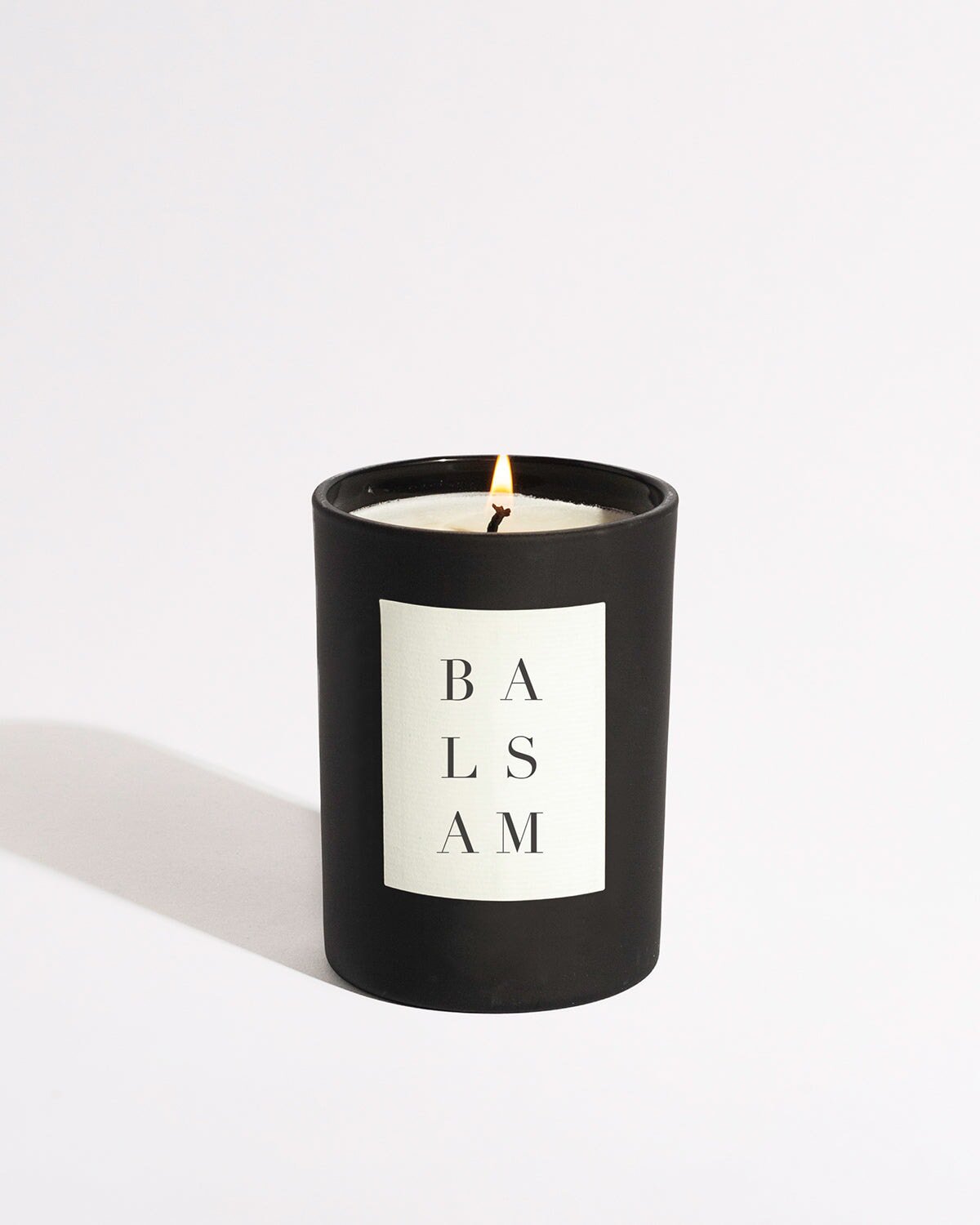 A lit candle, housed in a sleek black container with a white label that vertically reads "BALSAM," emits a warm glow. This eco-friendly Balsam Noir Candle by Brooklyn Candle Studio is crafted from soy wax, set against a plain white background.