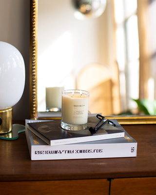 A lit Brooklyn Escapist Candle by Brooklyn Candle Studio in a glass jar sits on top of a stack of books, including one titled "Brooklyn." The books are on a wooden table in front of a mirror. A modern table lamp with a rounded white shade and a gold base is situated to the left of the books.