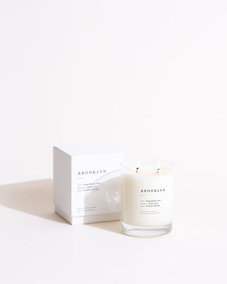 A minimalist white candle in a clear glass holder is placed in front of a white box. Both are labeled "Brooklyn Escapist Candle by Brooklyn Candle Studio." The setup, featuring eco-friendly candles, is against a plain white background, creating a clean and elegant presentation.