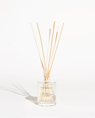 The Catskills Reed Diffuser by Brooklyn Candle Studio is a clear glass bottle labeled "Catskills," featuring light-colored reed sticks that gracefully extend from its neck. Filled with a transparent botanical aroma, this elegant setup rests against a plain white background, casting a faint shadow—an ideal addition to infuse serenity into your home office.