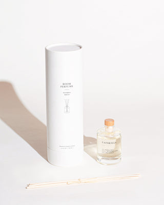 A minimalist product setup featuring the Catskills Reed Diffuser by Brooklyn Candle Studio, perfect for your home office. The image showcases a white cylindrical box labeled "ROOM PERFUME," a clear glass bottle with a wooden cap labeled "CATSKILLS" filled with liquid, and a bundle of white reed sticks that provide a botanical aroma.