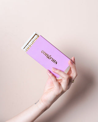 A hand with a gold ring holds the CONGRATS Lilac Long Matches by Brooklyn Candle Studio, featuring "congrats" in gold lettering. The lilac matchbox is partially open, revealing white-tipped XL matches inside. Perfect for special occasions, the background is a soft, light beige color.