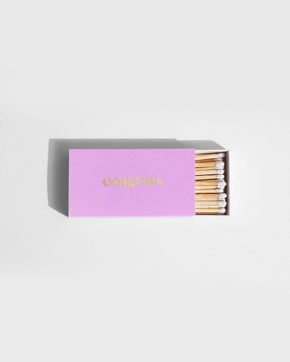 The CONGRATS Lilac Long Matches by Brooklyn Candle Studio feature a chic matchbox with a purple cover adorned with the word "congrats" in shimmering gold letters. The partially open box showcases the white-tipped XL matches inside, making it ideal for special occasions. The stylish design is highlighted against a simple white background.