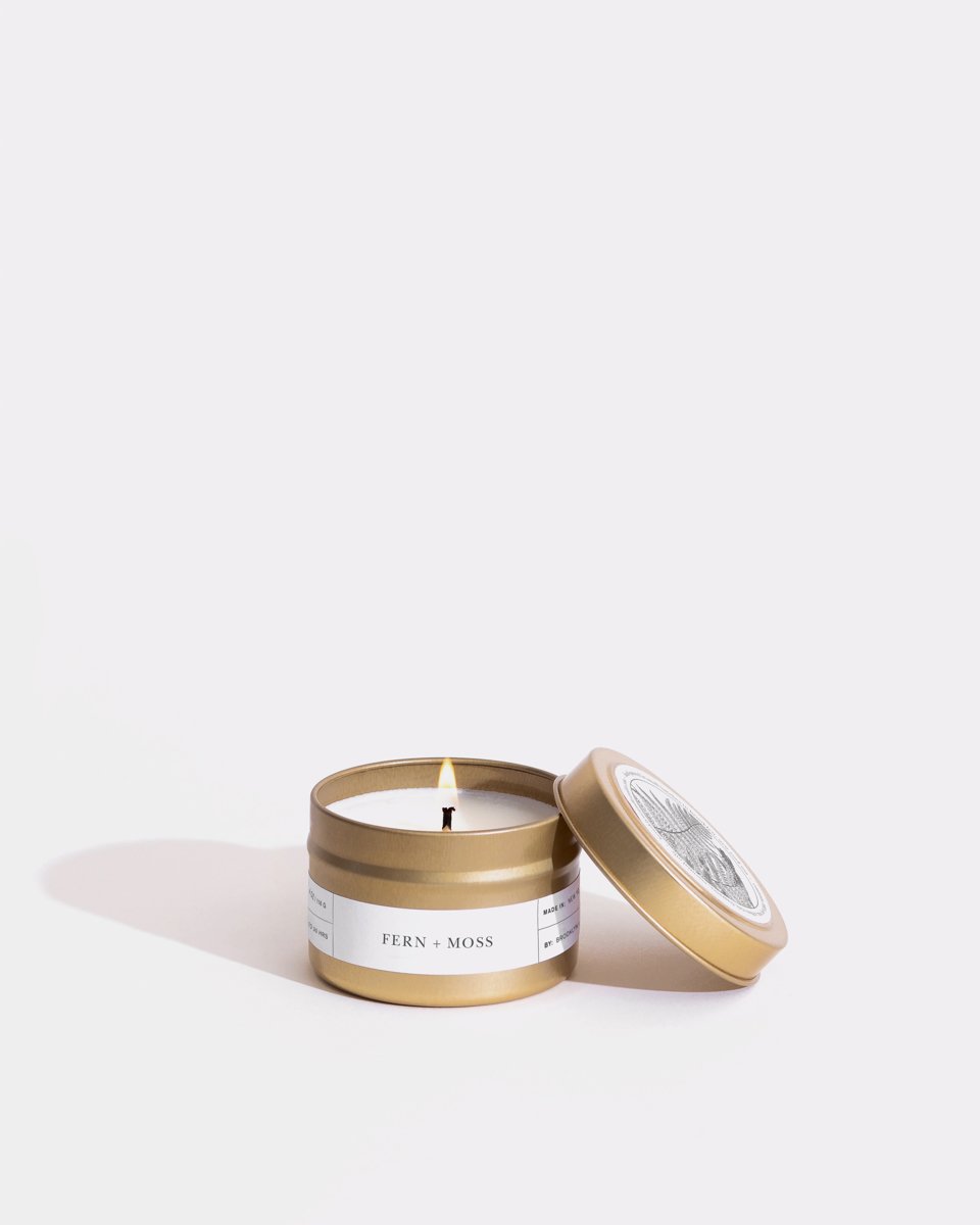 A small gold tin labeled "Fern + Moss Gold Travel Candle by Brooklyn Candle Studio" contains a lit soy wax candle. The tin's lid is off and leaning against the tin on a light purple background. The label is white with black text, and the candle's wick is slightly burnt. Shadows cast to the left evoke an eco-friendly ambiance with a Fern Canyon fragrance.