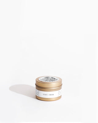 A small cylindrical tin container with a gold lid and a white label reading "Fern + Moss Gold Travel Candle by Brooklyn Candle Studio." Featuring a black and white illustration of foliage, the eco-friendly candle casts a soft shadow on a white background.