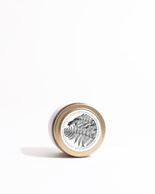 A small tin can with a gold lid rests on its side against a white background. The label on the lid features a black and white illustration of detailed fern leaves along with text around the border, promoting the Fern + Moss Gold Travel Candle by Brooklyn Candle Studio fragrance. The image is minimalist with soft shadows.