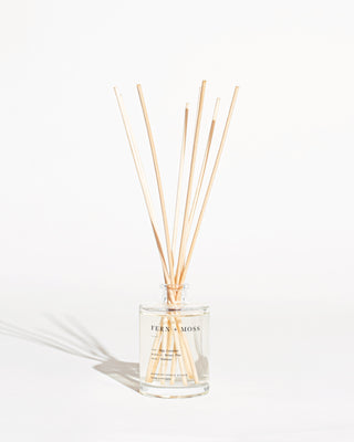 A clear glass bottle labeled "Fern + Moss Reed Diffuser by Brooklyn Candle Studio" holds several long, thin reed sticks that are fanned out. The bottle appears to contain a transparent liquid, likely a botanical aroma fragrance oil, and it is set against a plain white background.