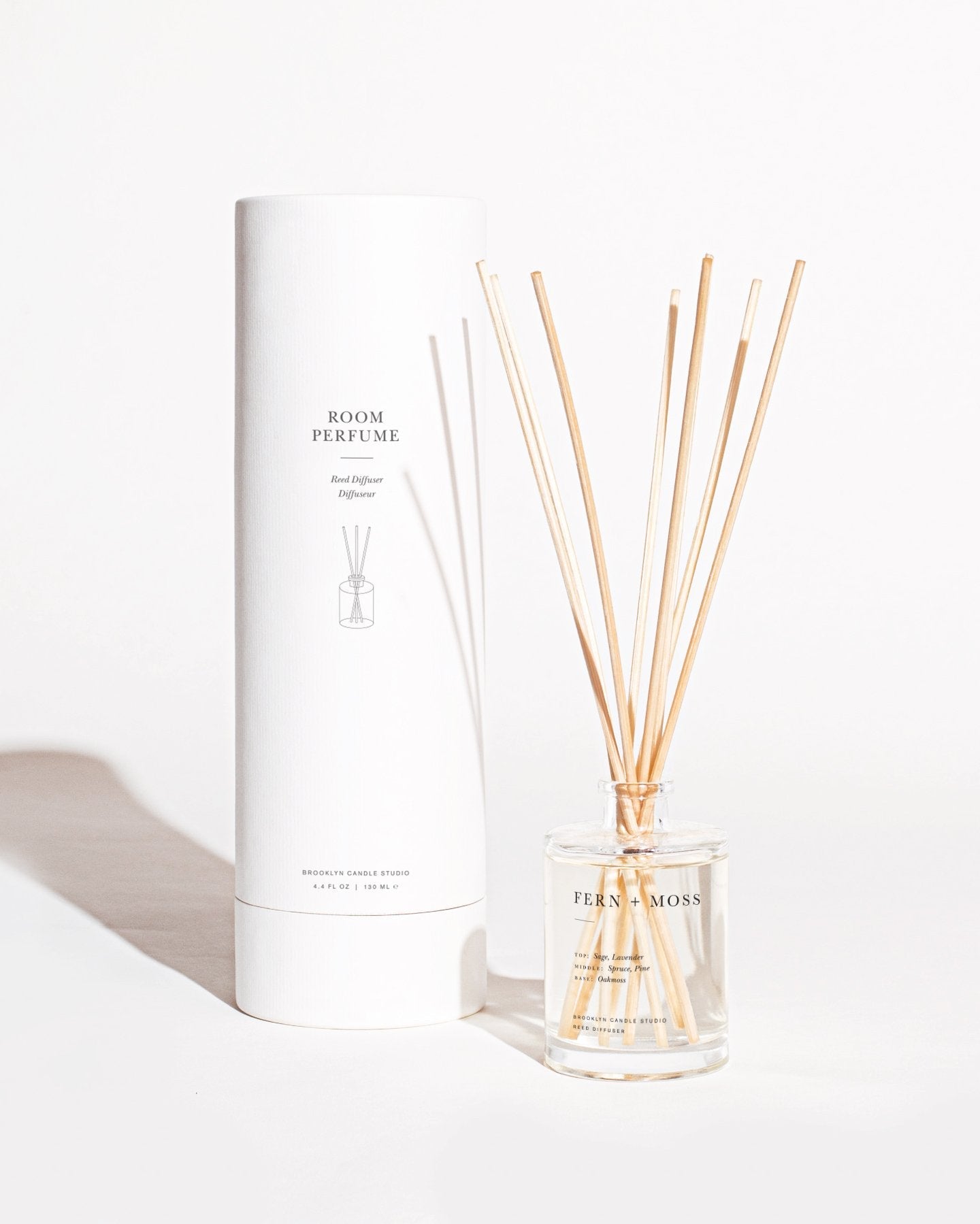 A clear glass bottle of Fern + Moss Reed Diffuser by Brooklyn Candle Studio with several natural-colored reeds sticking out of it. The botanical aroma infuses the space, and the bottle is placed next to a tall white cylindrical box labeled "Room Perfume" on a white background.