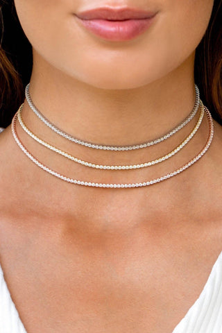 A close-up image showcases a person wearing three delicate Thin Tennis Chokers by By Adina Eden in silver, gold plated sterling silver, and rose gold hues. These necklaces, adorned with dazzling CZ stones, are neatly layered on the person's neck, perfectly complementing their light-colored, textured top. Their lips are slightly parted and blonde hair partially visible.