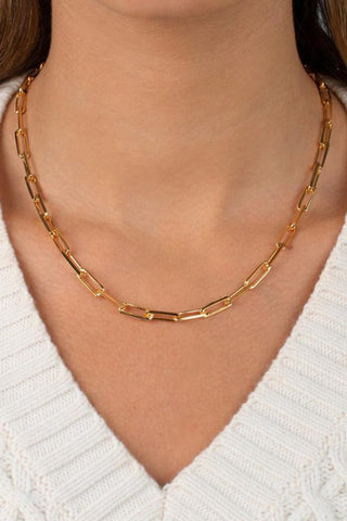 Close-up of a person wearing the Large Paperclip Link Necklace by Adina Eden. The necklace, characterized by its rectangular links, is styled over a white, cable-knit sweater with a v-neckline. The person's hair partially cascades down the sides. This versatile accessory adds elegance to any outfit.