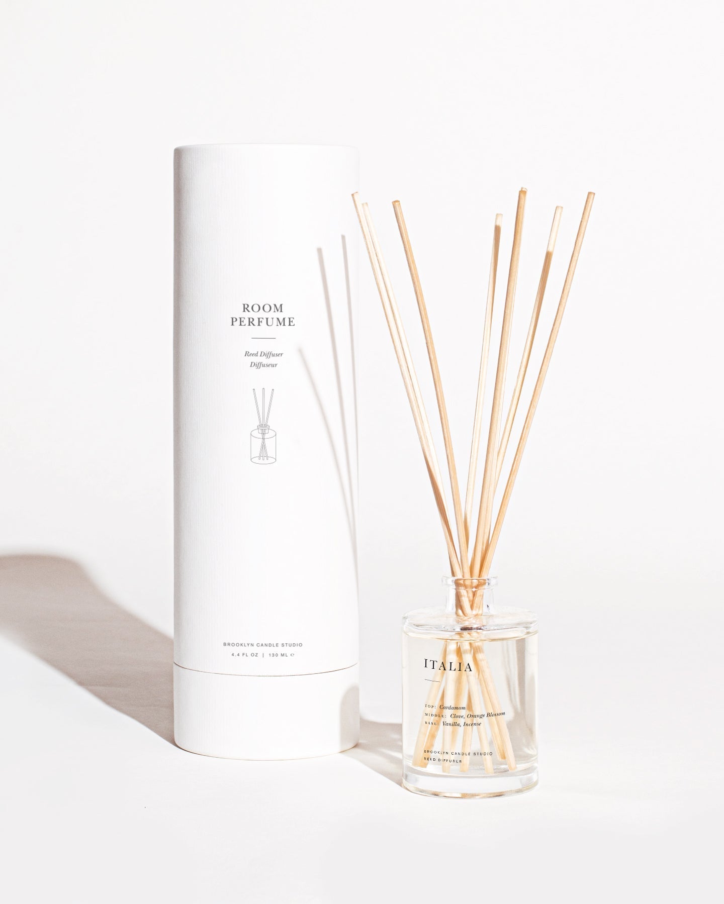 A room perfume set featuring a cylindrical white box labeled "Room Perfume" next to an open bottle of diffuser fragrance with several wooden reeds sticking out. The bottle, emblazoned with "ITALIA" in uppercase letters, emits a botanical aroma with hints of exotic spices. The background is white. This is the Italia Reed Diffuser by Brooklyn Candle Studio.