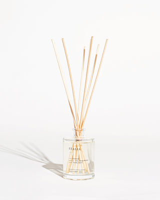 A clear glass diffuser bottle labeled "ITALIA" contains a light-colored liquid and several natural reed sticks emerging from its neck, fanned out and casting soft shadows. Infused with an exotic spices aroma, the background is white, creating a minimalist and clean appearance. The product is the Italia Reed Diffuser by Brooklyn Candle Studio.