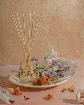 A glass bottle of Italia Reed Diffuser by Brooklyn Candle Studio with multiple reed sticks is placed on a beige plate. Next to it are broken pieces of cookies wrapped in translucent green tissue paper and purple flowers. The background and surface are pinkish-beige tiles. Scattered petals surround the setup, exuding a botanical aroma.