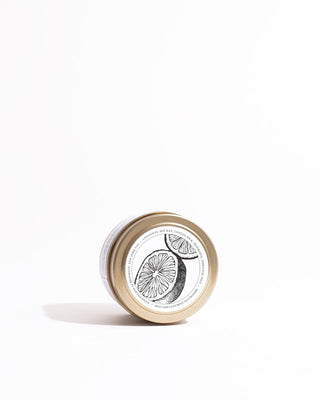 The eco-friendly Japanese Citrus Gold Travel Candle by Brooklyn Candle Studio, housed in a small cylindrical container with a metallic lid and featuring an illustration of two citrus slices, is lying on its side against a white background.