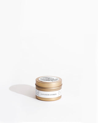 A small, gold-colored tin container labeled "Japanese Citrus Gold Travel Candle by Brooklyn Candle Studio" with a white and black lid design is placed against a plain white background. This eco-friendly candle casts a soft shadow to the left.