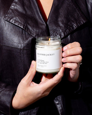 A person wearing a dark leather jacket is holding a lit candle labeled "Leather Jacket Minimalist Candle by Brooklyn Candle Studio." The label also displays scent notes including Saffron, Jasmine, Amber, Birch, and Cedar. Crafted from eco-friendly soy wax candles, the person's hands are shown with a ring on one finger.