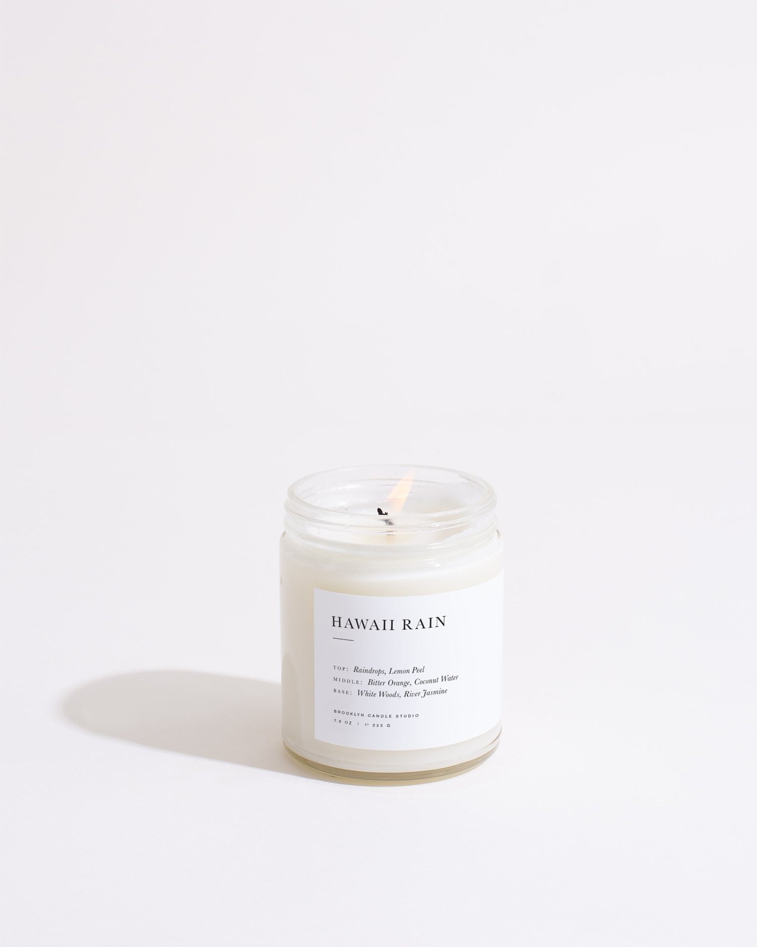 A lit white candle in a clear glass jar is centered against a light background. The label on the jar reads "Hawaii Rain Minimalist Candle" by Brooklyn Candle Studio, featuring scent notes of raindrops, lemon peel, and white tea. Made from soy wax, the flame casts a subtle glow and shadow around the candle.