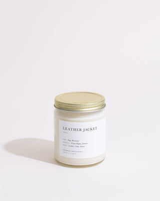 A medium-sized white jar candle with a golden lid is displayed against a plain white background. The label on the jar reads "LEATHER JACKET" and highlights scent notes of sage and rosemary, among other ingredients. This 11 oz eco-friendly candle, named Leather Jacket Minimalist Candle by Brooklyn Candle Studio, uses soy wax for a cleaner burn.