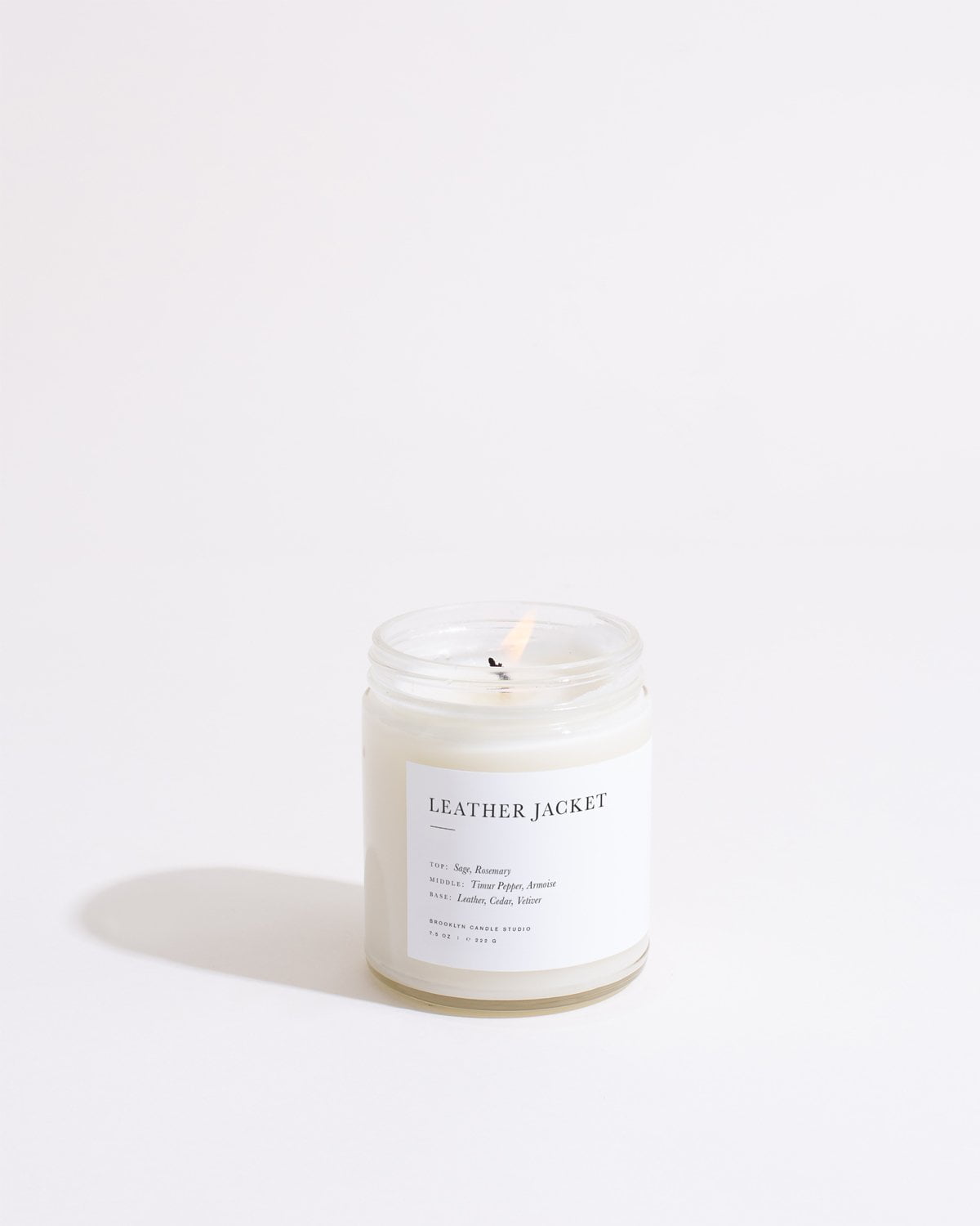 A lit candle in a transparent glass jar labeled "LEATHER JACKET" by Brooklyn Candle Studio is placed on a white surface. The label lists notes of various scents: "Soy, Fairmount, Burch, Honey, Jasmine, Leather, Mint, Lime." This eco-friendly Leather Jacket Minimalist Candle throws a soft glow with its shadow visible.