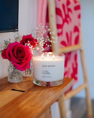 A lit white candle labeled "Love Potion Maximalist 3-Wick Candle by Brooklyn Candle Studio," featuring premium fragrance oils and multiple small flames, sits on a wooden surface next to a vase of red roses and baby's breath. In the background are pink-red patterned fabrics draped over a wooden ladder. This eco-friendly candle is crafted by Brooklyn Candle Studio.