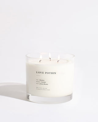 A white candle with three wicks in a clear glass container. The label reads "Love Potion Maximalist 3-Wick Candle by Brooklyn Candle Studio" with notes of Muskrose, Amber, and Jasmine Blossom beneath. Crafted by Brooklyn Candle Studio using premium fragrance oils, the eco-friendly candle sits on a plain, light-colored background, shadow to the left.