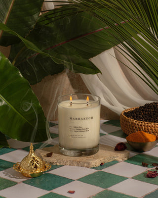 A lit Marrakech Escapist Candle by Brooklyn Candle Studio with two flames sits on a stone in a lush setting adorned with green leaves and flowing white fabric in the background. To the right, a wicker bowl holds black peppercorns and a dried rose. A decorative gold object graces the foreground. This eco-friendly soy wax creation is cruelty-free.