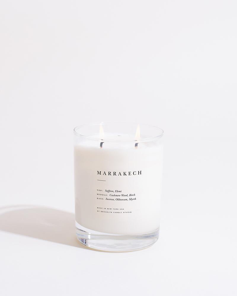 A lit Marrakech Escapist Candle by Brooklyn Candle Studio, nestled in a clear glass holder and made of eco-friendly soy wax, sits surrounded by plain white. The candle, with two small flames flickering from its wicks, carries a sophisticated fragrance blend described on its label: notes of saffron, vetiver, incense, amber, and musk.