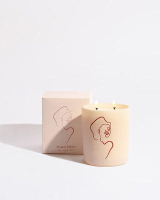 A beige candle with two wicks, featuring a minimalist red line drawing of a face by Allison Kunath on its side. Behind it is the matching square box with the same design and text that reads "Mojave Embers - Allison Kunath Artist Edition Candle by Brooklyn Candle Studio" along with a description. All items are set against a plain light background, embodying a minimalist aesthetic.