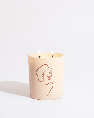 A light pink candle featuring minimalist line art by Allison Kunath, depicting a human face on its surface. The Mojave Embers - Allison Kunath Artist Edition Candle by Brooklyn Candle Studio is lit with two flames and set against a plain white background, perfectly capturing the minimalist aesthetic.