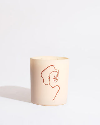 A beige Mojave Embers - Allison Kunath Artist Edition Candle by Brooklyn Candle Studio in a minimalist holder, adorned with a simple red line drawing of a person's face by Allison Kunath on the front. The background is plain white, giving the image a clean and modern look that perfectly captures the minimalist aesthetic.