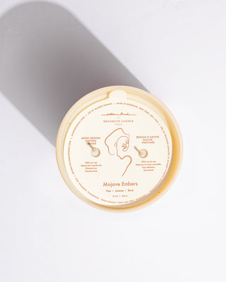 A top-down view of a Mojave Embers - Allison Kunath Artist Edition Candle by Brooklyn Candle Studio. The cream-colored candle, featuring two wicks, showcases minimalist, artistic line drawings by Allison Kunath on its label. The text indicates a scent profile of pine, incense, and birch.