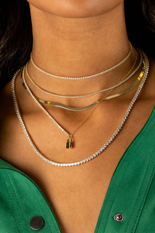 A close-up of a person wearing multiple layered necklaces features the Thin Tennis Choker by By Adina Eden, adorned with dazzling CZ stones. The ensemble includes a gold-plated sterling silver chain and a diamond-studded chain with a small padlock pendant. The person is dressed in a green button-up shirt, slightly open at the collar.