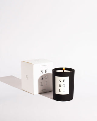 A lit **Neroli Noir Candle by Brooklyn Candle Studio** sits next to its white packaging box on a white surface. The candle and box both display the word "Neroli" in minimalist, uppercase letters. Made with premium fragrance oils, the candle's flame casts a gentle shadow, embodying eco-friendly elegance.