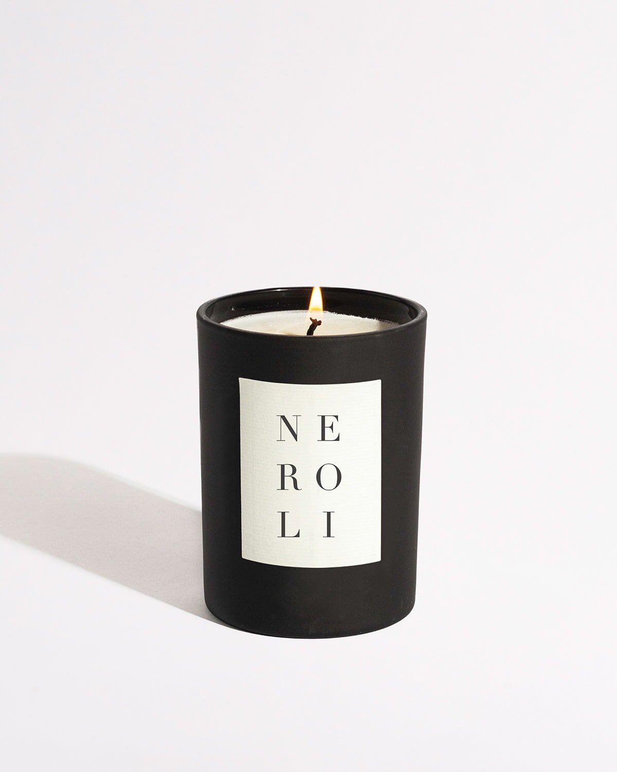 A black Neroli Noir Candle by Brooklyn Candle Studio, adorned with a white label featuring the word "Neroli" written vertically, is lit and casting shadows on a white background. This candle is crafted with premium fragrance oils for an elevated experience.