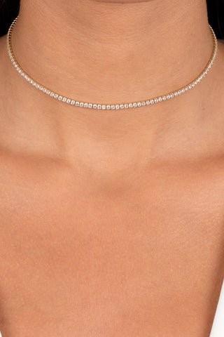 A close-up shot captures a person's neck adorned with the Thin Tennis Choker by By Adina Eden, featuring dazzling CZ stones set in gold-plated sterling silver. The individual is dressed in a white top with a V-shaped neckline. The warm skin tone and neutral background emphasize the exquisite jewelry piece.