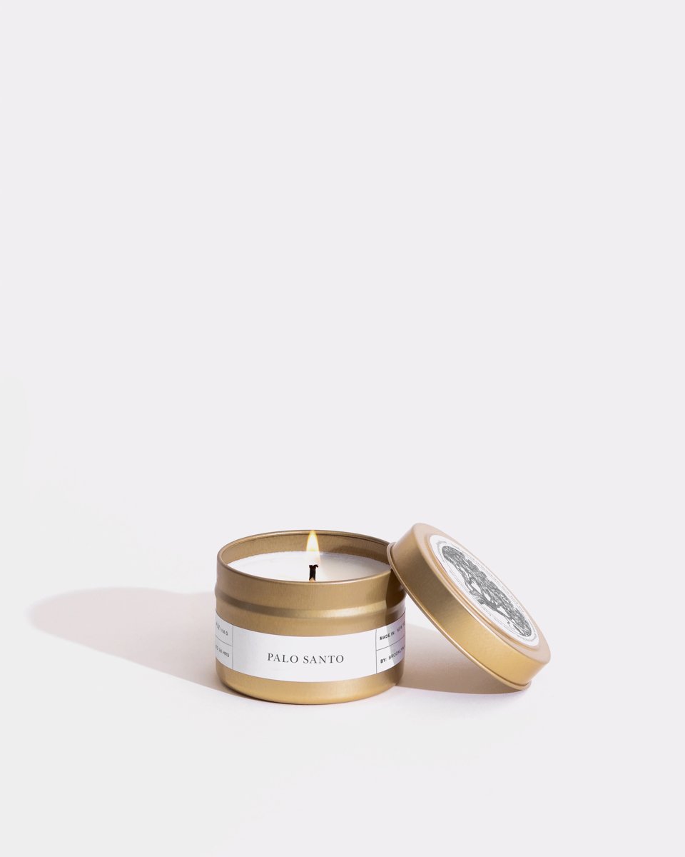 A small, open Palo Santo Gold Travel Candle by Brooklyn Candle Studio with a wooden wick. The eco-friendly gold tin, crafted from soy wax, features a white label adorned with black text and a detailed circular design on the lid, which is leaning against the side of the container. The background is plain white.