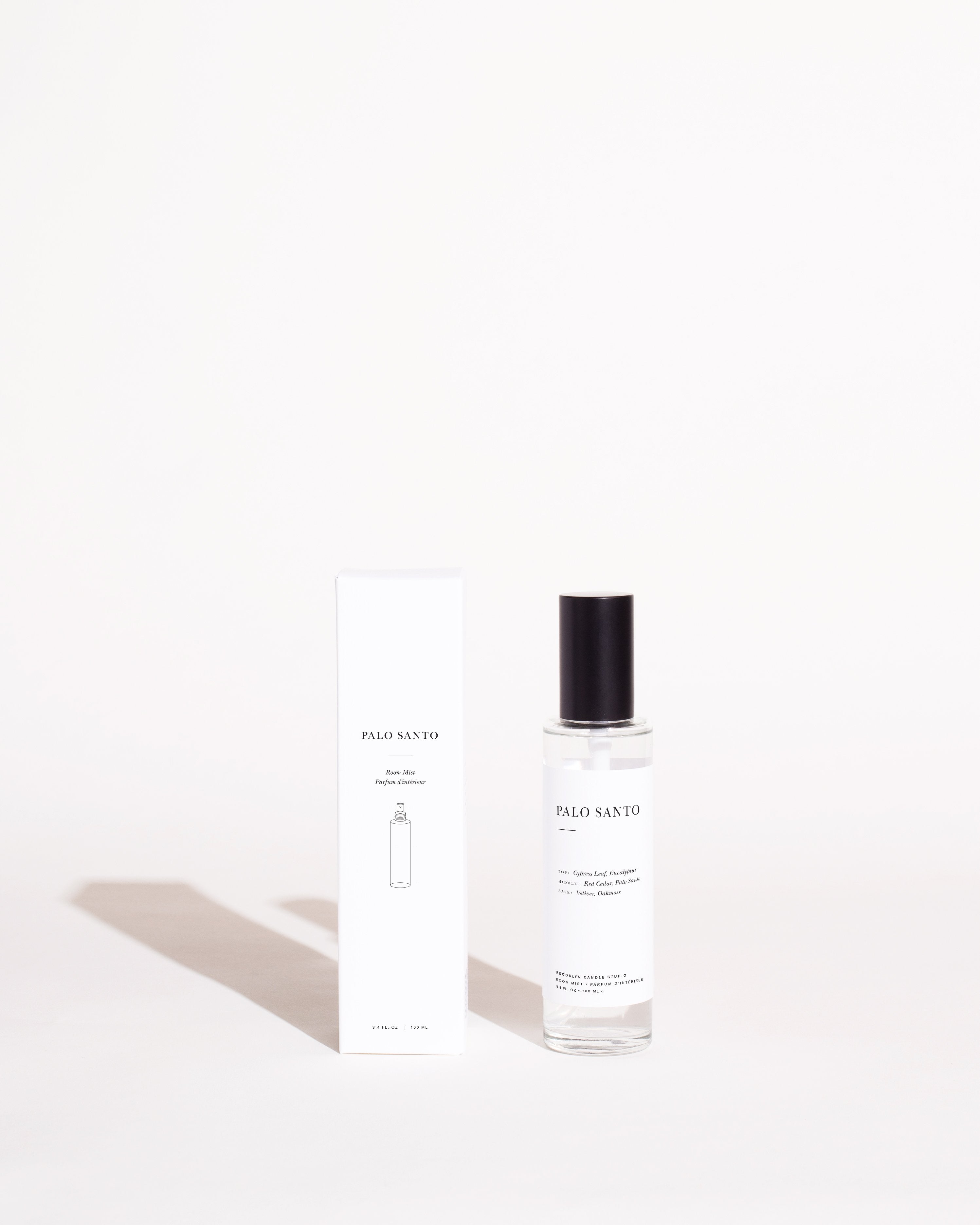 A minimalist photo featuring a Palo Santo Room Mist by Brooklyn Candle Studio bottle with a black cap next to its corresponding white, rectangular packaging box. Both the bottle and the box display the product name and descriptions in a simple, elegant font against a light background, emphasizing its essential oils blend.