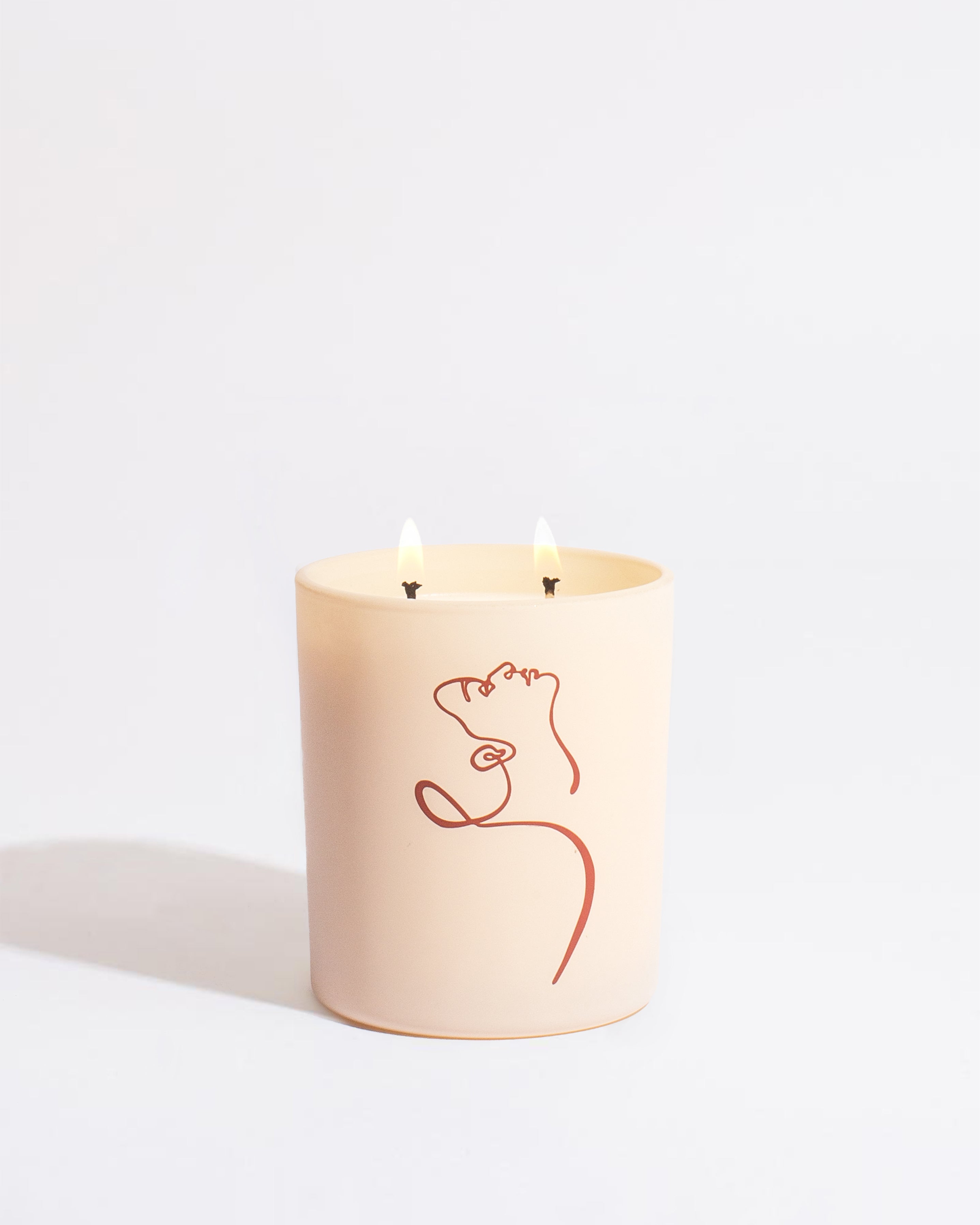A beige candle with two wicks burning softly from the Petrichor - Allison Kunath Artist Edition Candle by Brooklyn Candle Studio sits elegantly. The candle holder features a minimalist red line drawing of a woman's face and upper body, all against a plain white background.