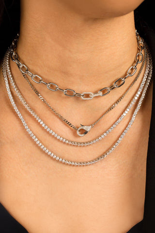 Close-up image of a person wearing multiple layered necklaces. The top necklace features a chunky chain, while the lower ones include the Three Prong Tennis Necklace by By Adina Eden and finer chains adorned with sparkling crystals. The gold-plated and sterling silver necklaces are elegantly stacked, creating a stylish, layered look.