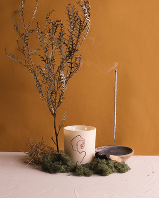 A minimalist setup featuring a potted dry branch, a yellow Mojave Embers - Allison Kunath Artist Edition Candle by Brooklyn Candle Studio, and a lit incense stick in a holder. The arrangement sits on a white surface with moss, against a solid mustard-yellow background.