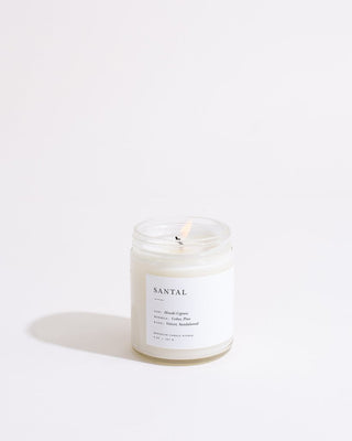 A lit Santal Minimalist Candle by Brooklyn Candle Studio, housed in a clear glass jar, casts a soft shadow to the left against a white, minimalist background. The label on the eco-friendly soy wax candle lists notes of Himalayan Cypress, Fig, Clary Sage, Lemon, Violet, and Sandalwood.