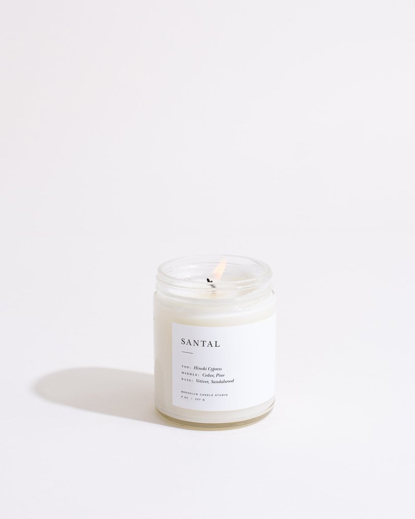 A lit Santal Minimalist Candle by Brooklyn Candle Studio, housed in a clear glass jar, casts a soft shadow to the left against a white, minimalist background. The label on the eco-friendly soy wax candle lists notes of Himalayan Cypress, Fig, Clary Sage, Lemon, Violet, and Sandalwood.