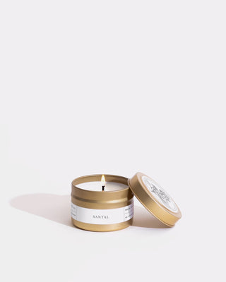 A small, open gold-colored tin container with a single wick soy wax candle labeled "Santal," featuring a white label with black text wrapped around the minimalist design. The lid rests diagonally beside it against a plain white background, showcasing the eco-friendly craftsmanship of Brooklyn Candle Studio’s Santal Gold Travel Candle.