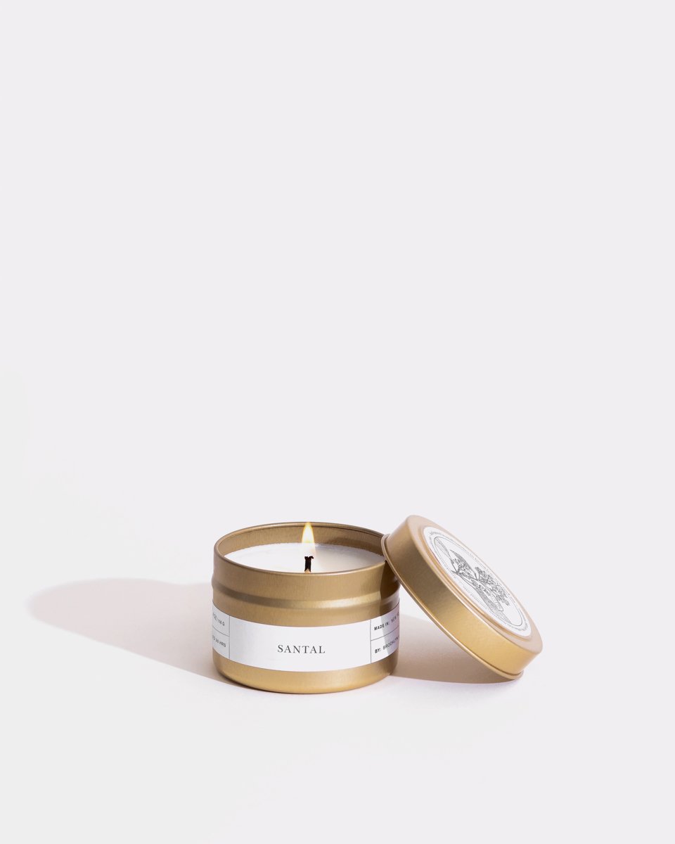 A small, open gold-colored tin container with a single wick soy wax candle labeled "Santal," featuring a white label with black text wrapped around the minimalist design. The lid rests diagonally beside it against a plain white background, showcasing the eco-friendly craftsmanship of Brooklyn Candle Studio’s Santal Gold Travel Candle.