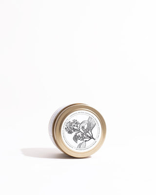 A small beige container with a gold lid lies on a white surface. It has a circular label featuring a monochrome botanical illustration of a plant with flowers and leaves. Text encircles the illustration, detailing the contents and ingredients of the Santal Gold Travel Candle by Brooklyn Candle Studio.